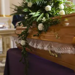 Drewniana trumna ozdobiona kwiatami i koronką, przygotowana do pogrzebu. W tle kościół.