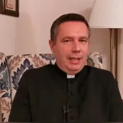 Ksiądz w czarnej sutannie siedzi w fotelu. Mówi o tym, ile za mszę w niedzielę, a za nim widać lampę i obrazy.