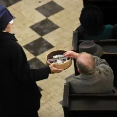Zakonnica w czarnym habicie trzyma koszyk z pieniędzmi, do którego starszy mężczyzna wkłada banknot.