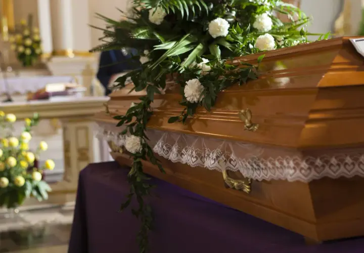 Drewniana trumna ozdobiona kwiatami i koronką, przygotowana do pogrzebu. W tle kościół.