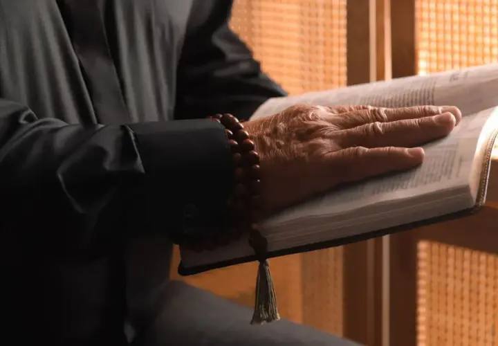 Dłoń starszego mężczyzny spoczywa na otwartej Biblii. Wokół nadgarstka ma drewniane paciorki. Gdzie jest pastor?