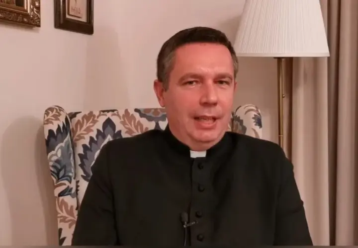 Ksiądz w czarnej sutannie siedzi w fotelu. Mówi o tym, ile za mszę w niedzielę, a za nim widać lampę i obrazy.