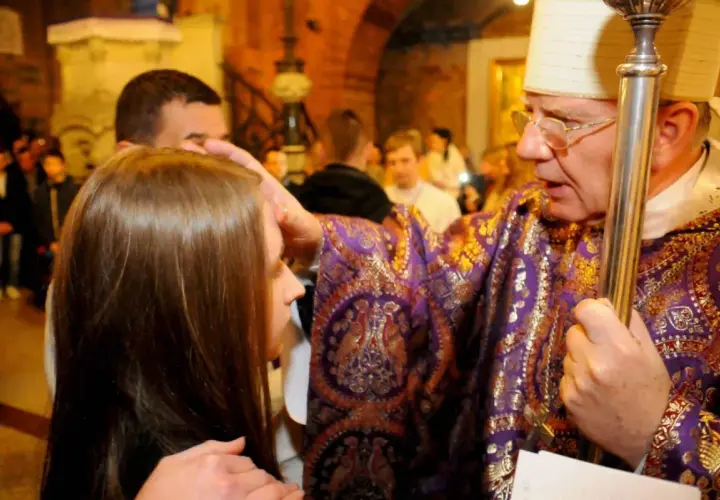 Biskup w bogato zdobionej szacie nakłada olej na czoło młodej kobiety. To moment bierzmowania dla dorosłych, symbol duchowego umocnienia.