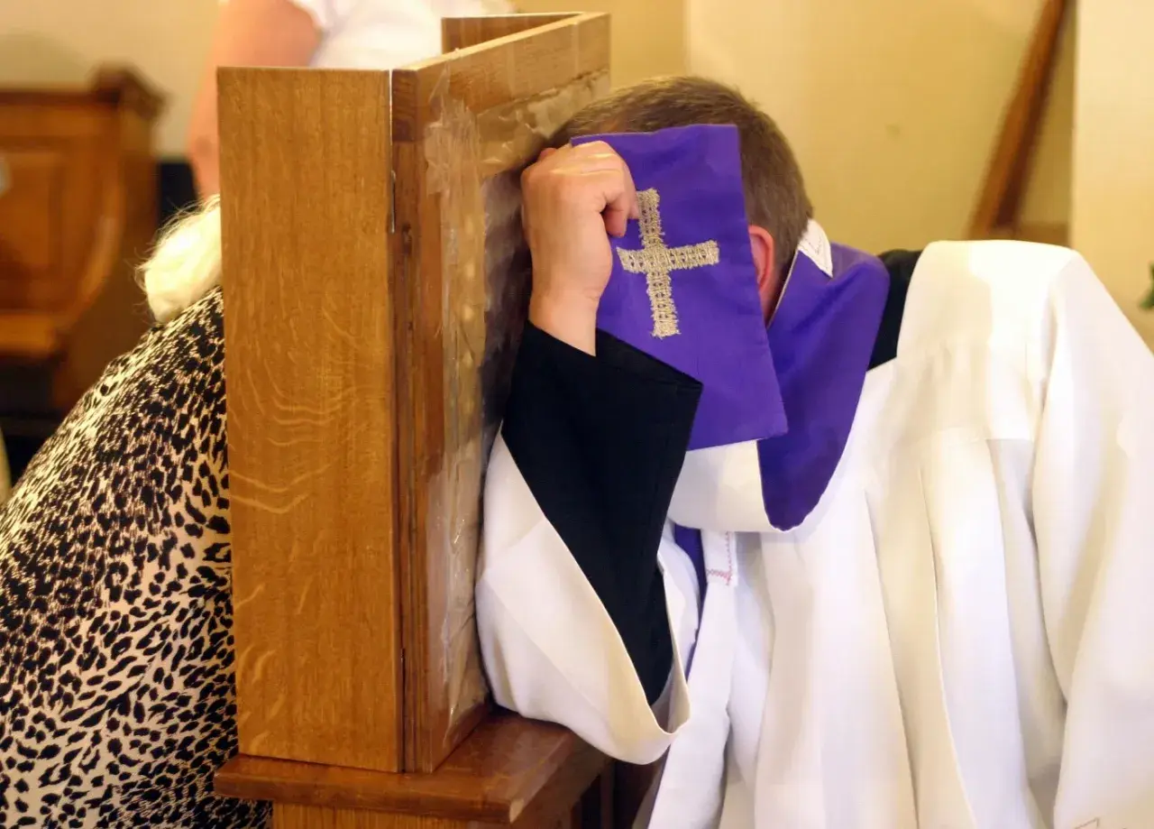 Spowiedź w konfesjonale. Czy nie pójście do kościoła z powodu choroby to grzech? Kapłan w białej albie i fioletowej stuły z krzyżem.