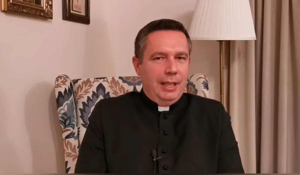 Ksiądz w czarnej sutannie siedzi w fotelu. Mówi o tym, ile za mszę w niedzielę, a za nim widać lampę i obrazy.