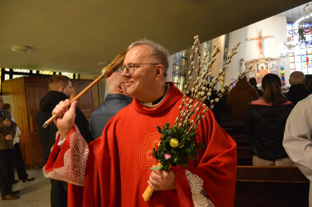 Ksiądz w czerwonym ornacie z baziami i kropidłem. W kościele trwa msza w Niedzielę Palmową.