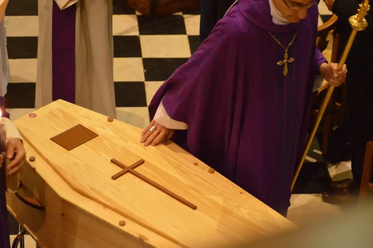Ksiądz w purpurowej szacie nad drewnianą trumną z krzyżem. Trwa msza za zmarłego, czas modlitwy i pożegnania.